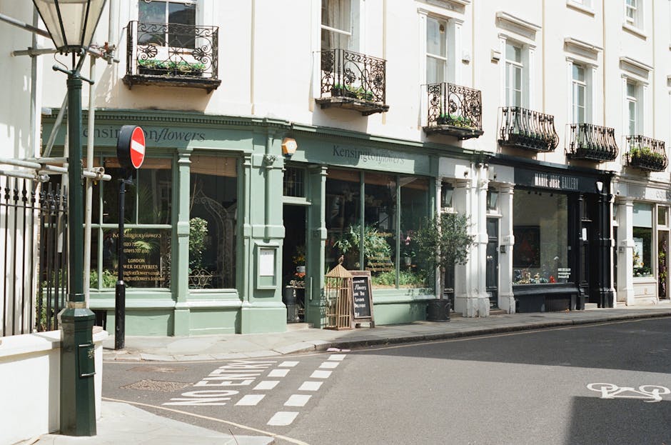 Exterior view of a charming flower shop in Barnes with a light green facade and large glass windows showcasing floral arrangements inside. The shop has decorative black wrought-iron balconies above, a