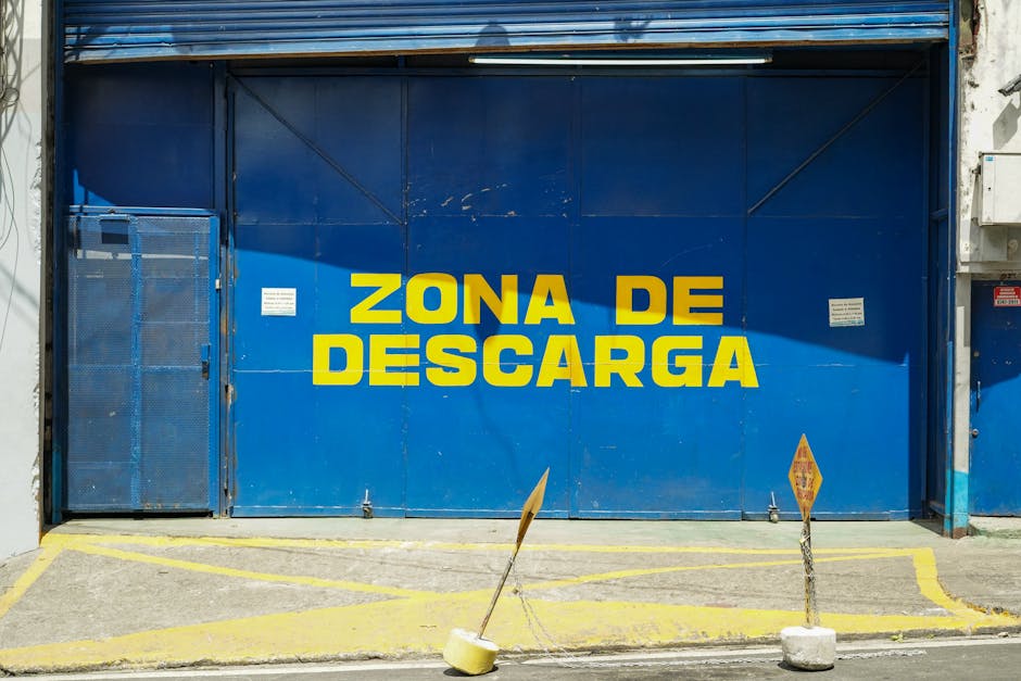 The image depicts a large blue metal garage door with bold yellow text reading 'ZONA DE DESCARGA,' indicating a loading zone. In front of the door, there are two waist-high yellow barriers with concre