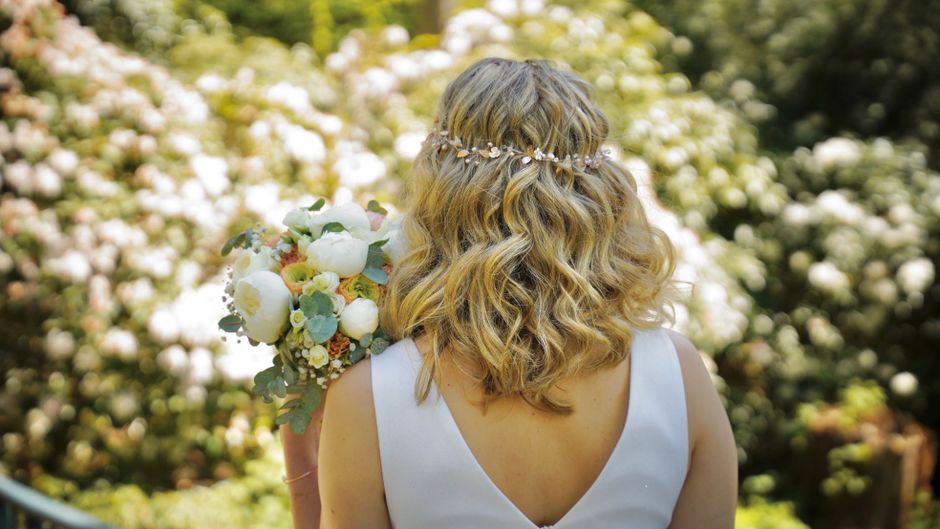 A woman with shoulder-length, wavy blonde hair, adorned with a delicate floral headpiece featuring small white flowers and greenery, is holding a lush bouquet of fresh flowers. The bouquet includes wh
